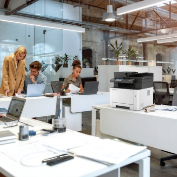 A modern white and black Kyocera multifunction printer sitting on a white desk in a bright, open-plan office, with colleagues working at desks in the background