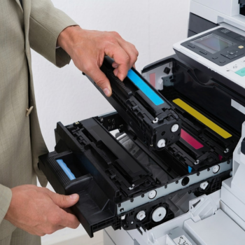 A person in a beige suit inserting a cyan toner cartridge into a large office colour laser printer.