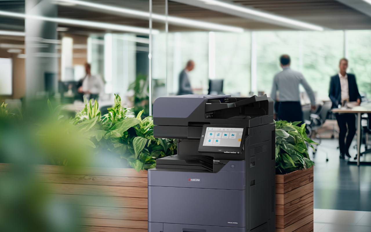 A grey multifunction office printer with a touch screen, positioned behind plants in a modern office environment.