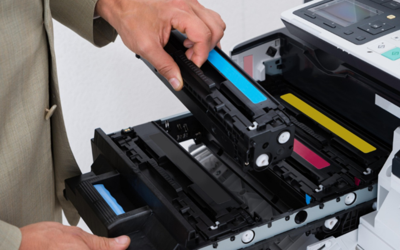 A person in a beige suit inserting a cyan toner cartridge into a large office color laser printer.